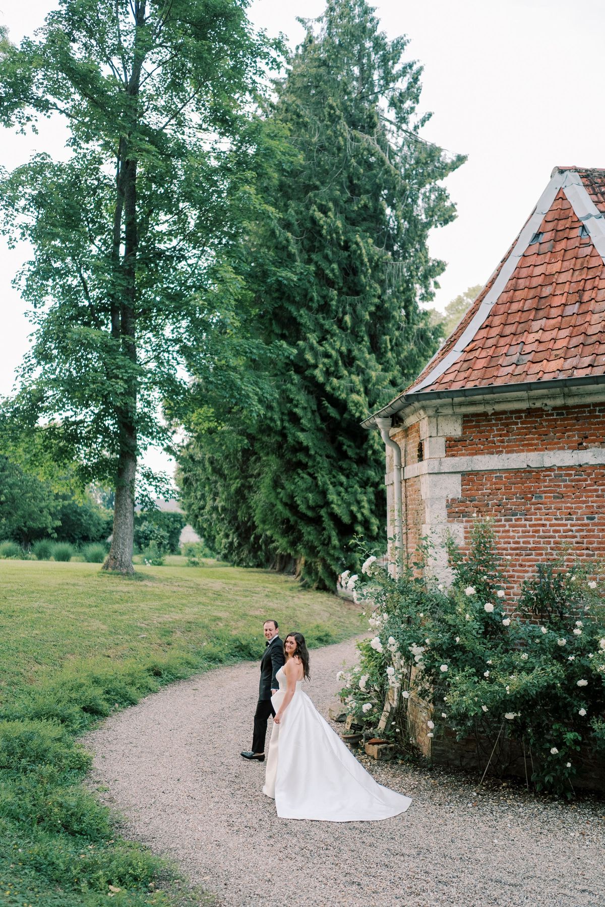 A gravel path through the parkland past a brick outbuilding planted with white roses