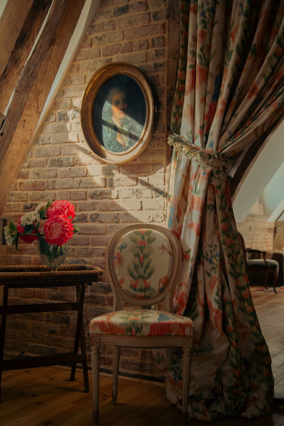 A corner of an attic room with a brick wall, an oval portrait and a floral upholstered chair