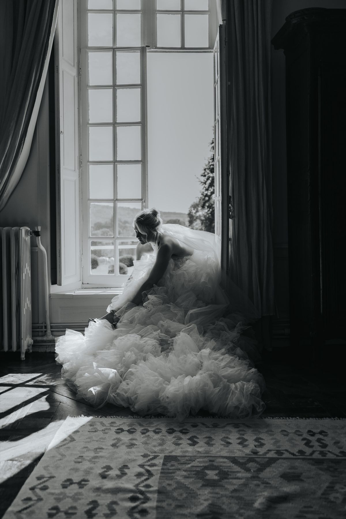 A bride by a tall window in a château room with a patterned rug