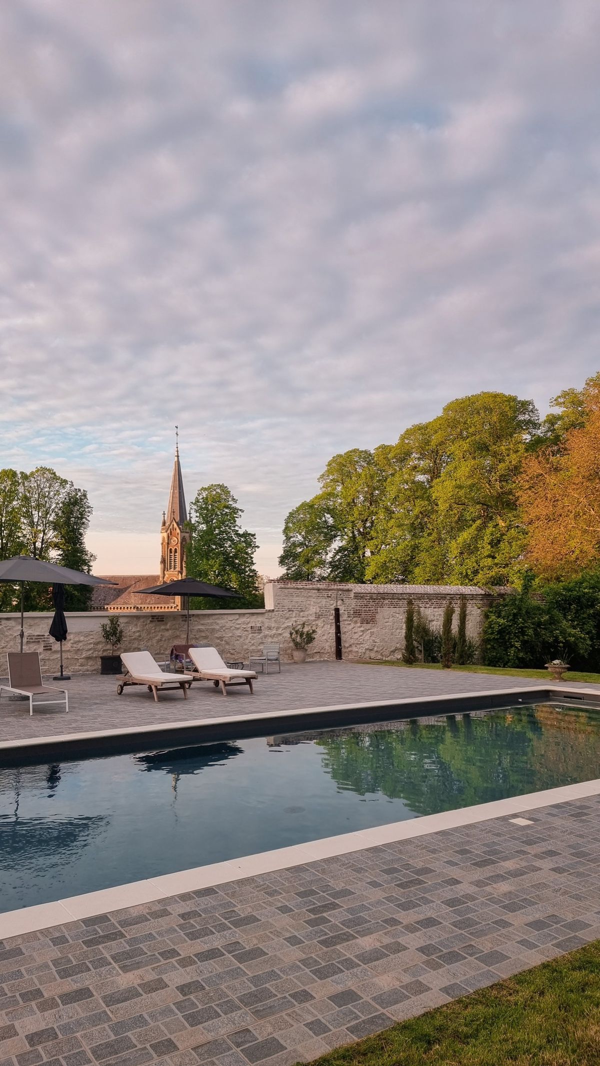 The pool terrace at dusk with the village church spire rising beyond the garden wall