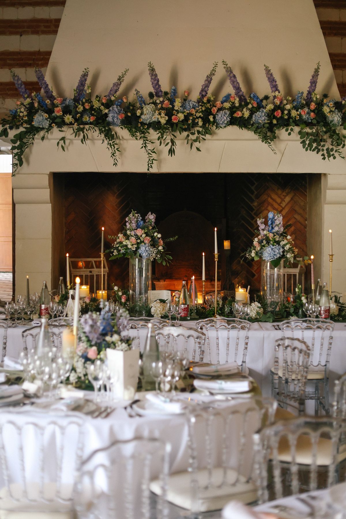 The reception room set for dinner beneath a large stone fireplace dressed with flowers