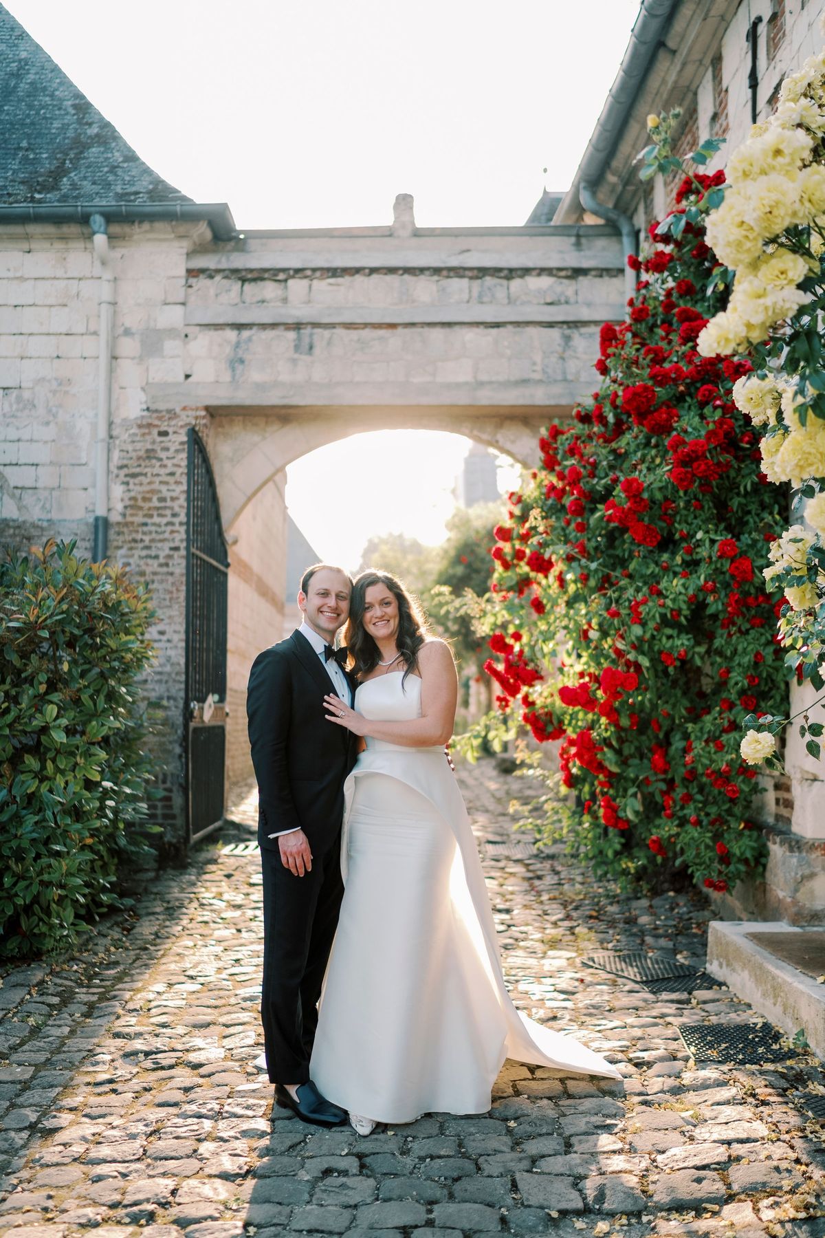 A stone archway heavy with climbing red and white roses over a cobbled path