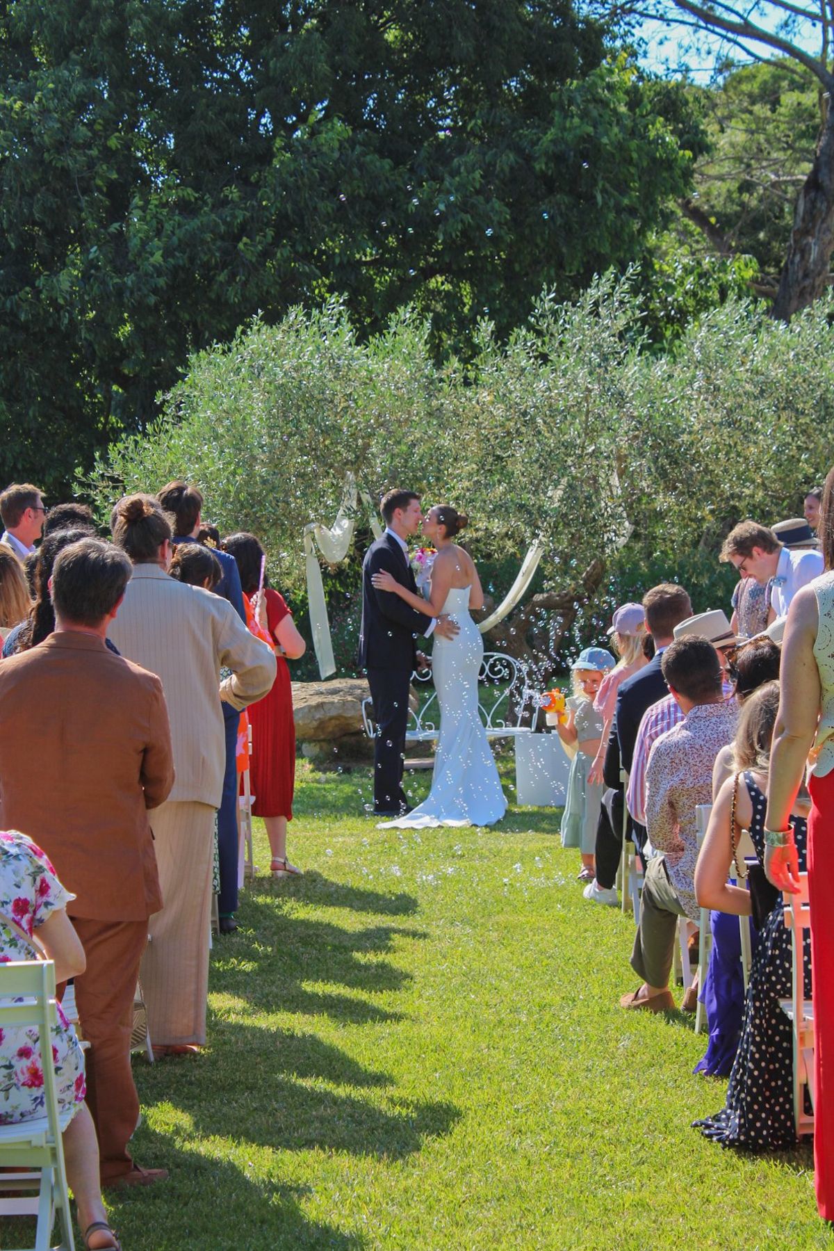 Guests standing either side of a grass aisle during a ceremony, the couple at the front beneath an olive tree hung with white drapes.