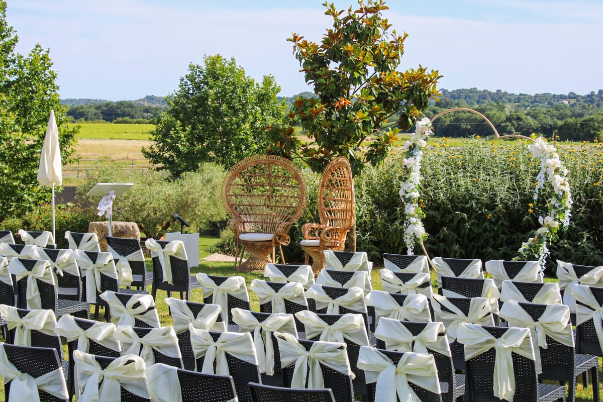 Rows of dark chairs tied with cream bows facing two wicker chairs and a circular floral arch, with a vineyard and wooded hills beyond.