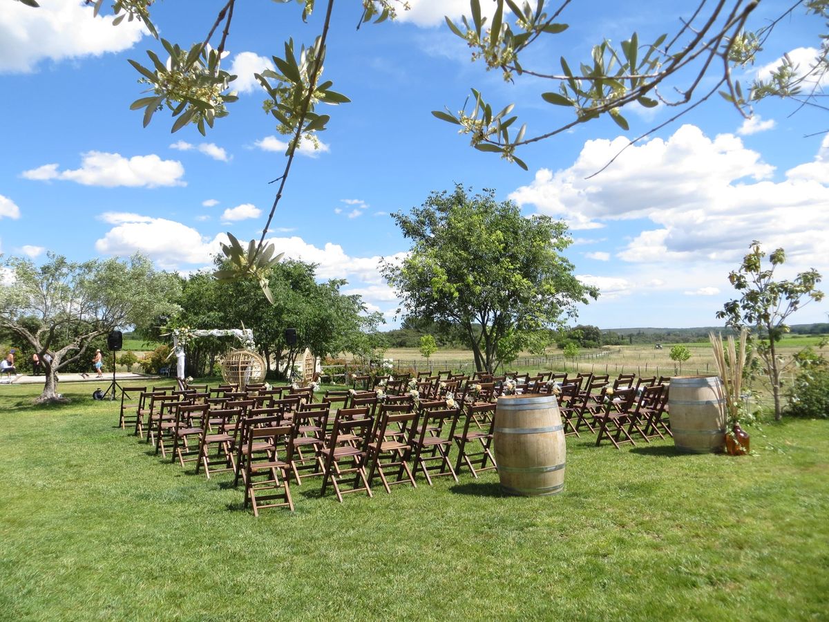 Rows of wooden folding chairs on a lawn facing a fabric and flower arch flanked by wine barrels, with open fields beyond a wire fence.