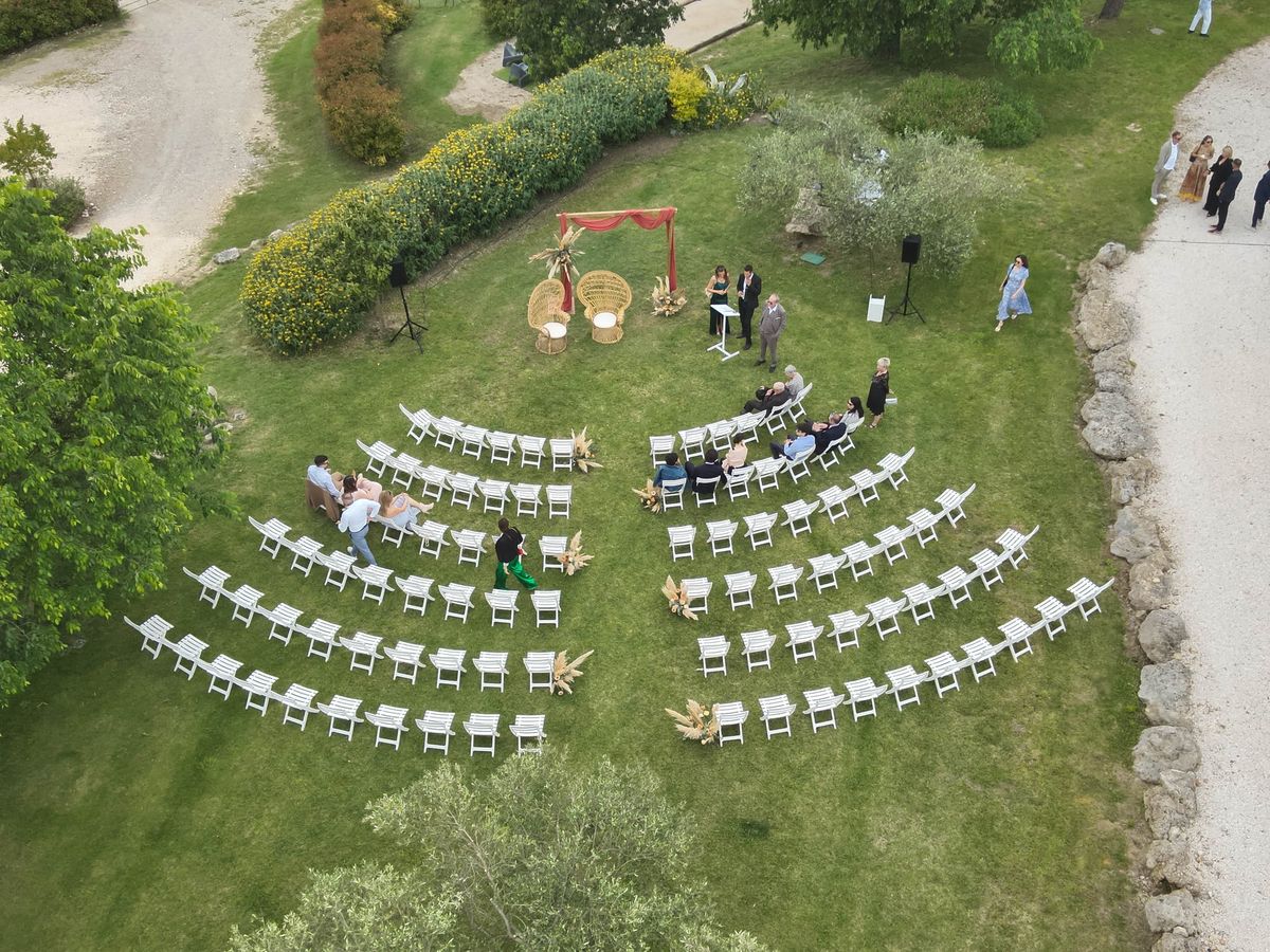 Aerial view of a lawn set with curved rows of white folding chairs facing a draped arch, with two wicker chairs and a lectern beside a flowering hedge.