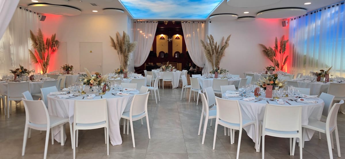 Indoor reception hall set with round tables in white linen, dried pampas arrangements and coloured uplighting, opening through to a second room.