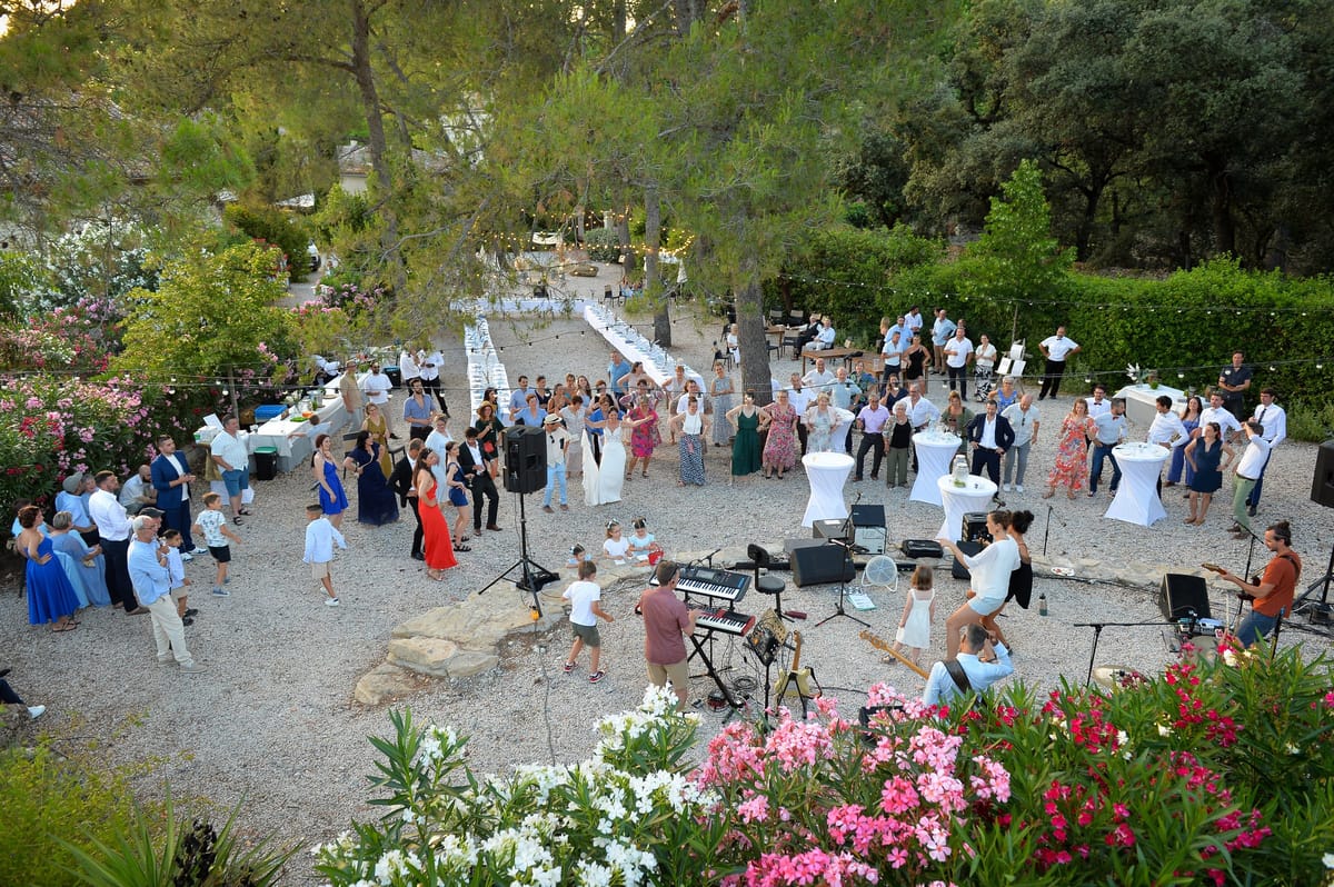 A cocktail reception on a gravel terrace with a band playing, guests at white poseur tables and oleander in flower along the edge.