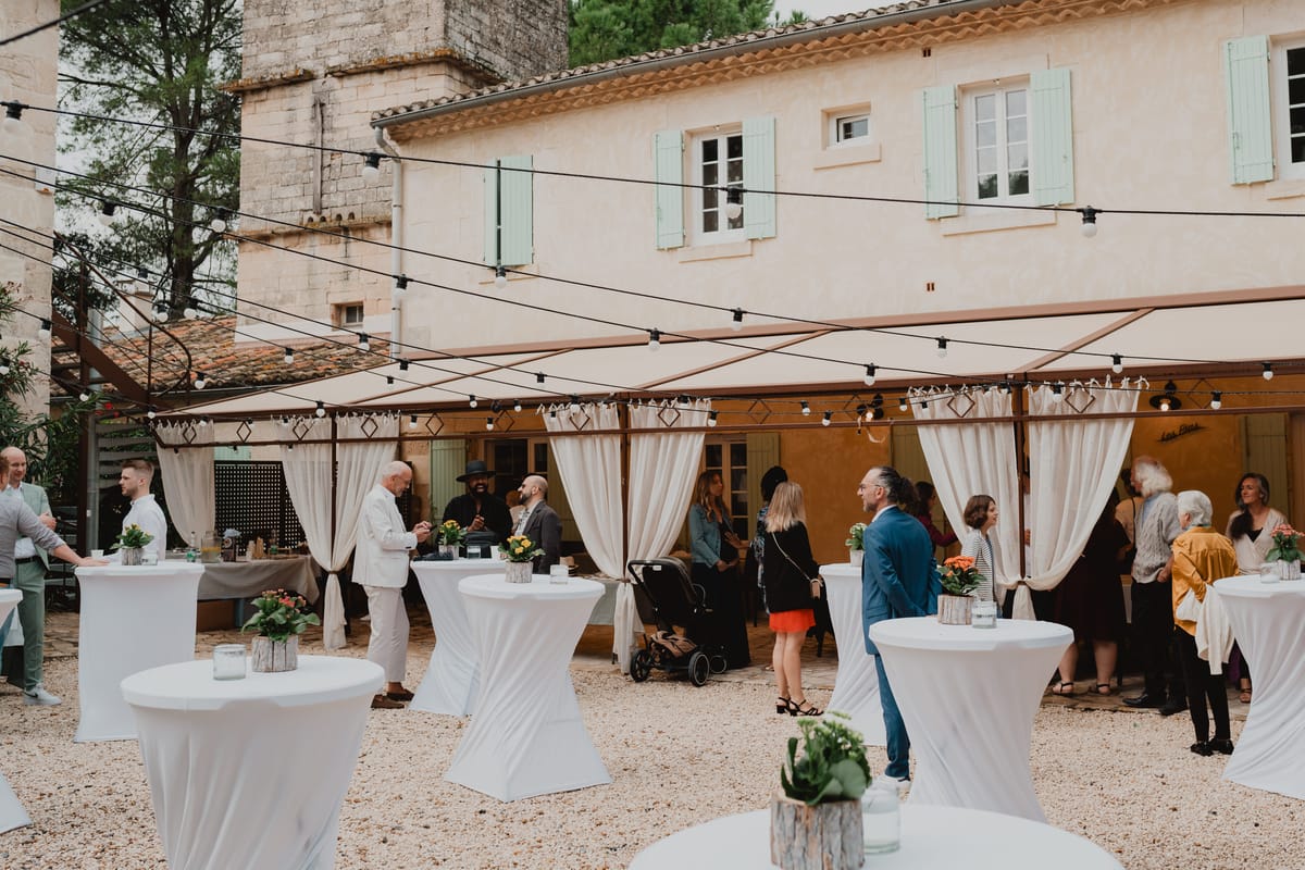 Cocktail reception with white high tables in a gravel courtyard beside a shuttered stone building, under awnings and festoon lights.