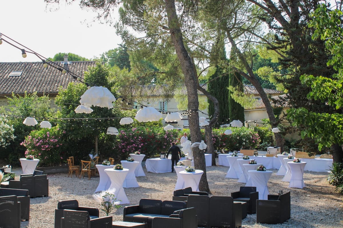 Gravel courtyard set for a drinks reception with white poseur tables, rattan lounge seating and fabric shades hung between the trees.