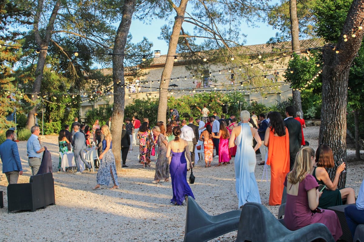 Guests at a drinks reception on the gravel beneath pine trees strung with festoon lights, the stone buildings behind.