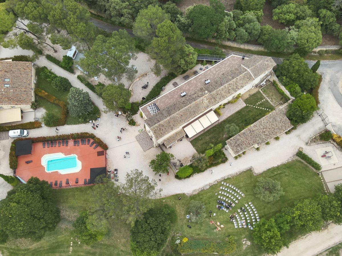 Aerial view of the estate showing a swimming pool on a tiled deck, long tiled buildings, gravel drives and a lawn set for a ceremony, surrounded by woodland.