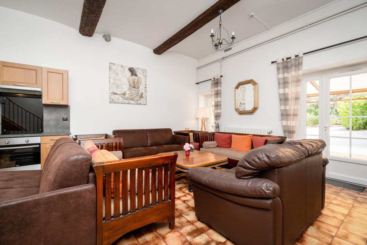 The living room of the Les Lauriers Roses gite, with brown leather sofas, exposed beams, a terracotta tiled floor and the kitchen beyond.