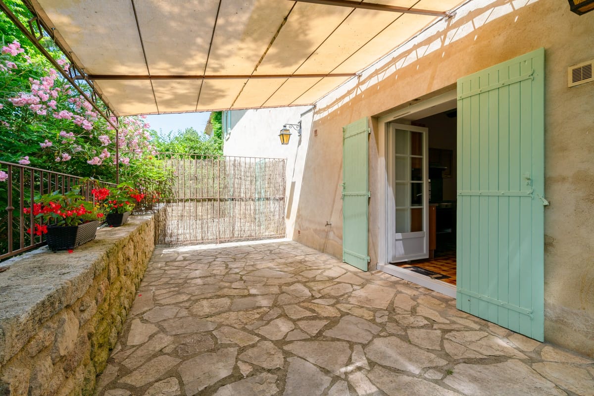 The stone-paved terrace of the Les Lauriers Roses gite, under a canvas awning beside pale green shutters, with potted geraniums and climbing roses.