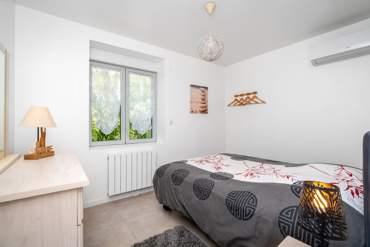A bedroom in the Les Daims gite with a double bed, a window looking onto greenery, a bedside lamp and a wall-mounted air conditioning unit.
