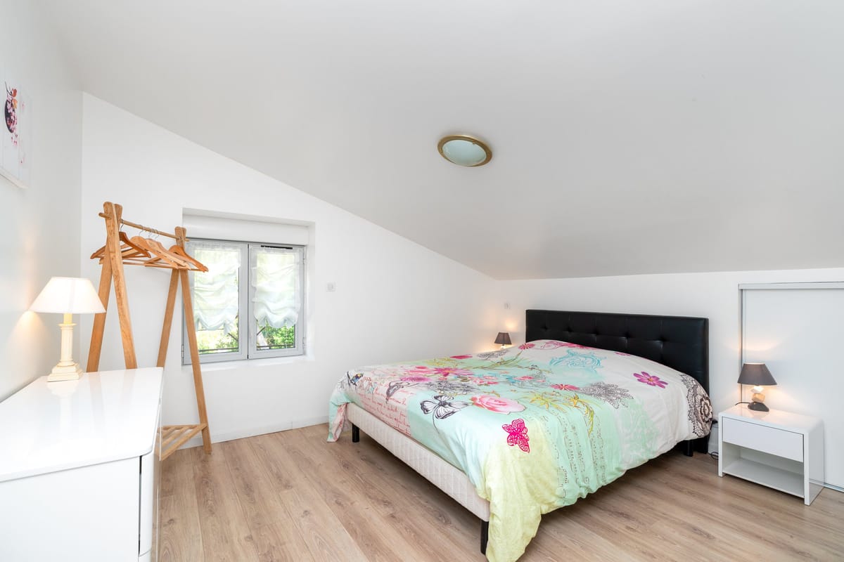 A bedroom in the Les Daims gite set under a sloping ceiling, with a double bed, a black upholstered headboard, a wooden clothes rail and a white chest of drawers.