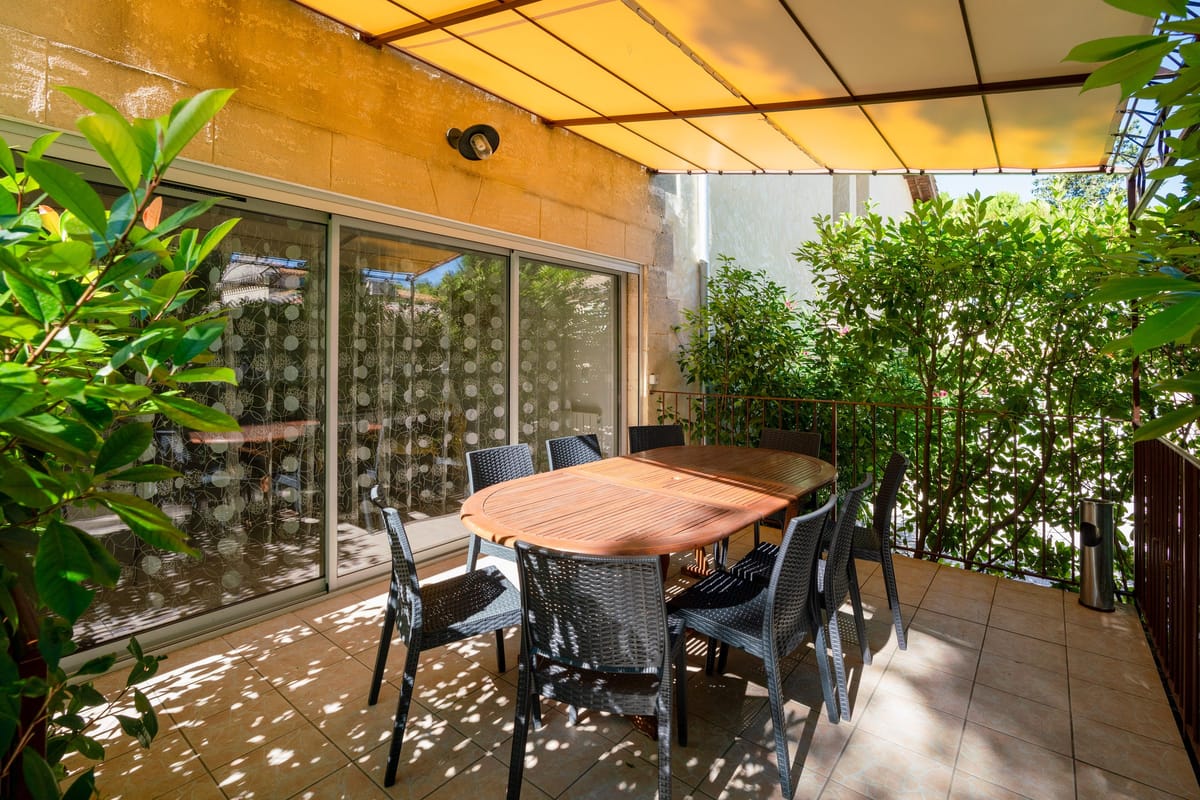 The enclosed entrance terrace of the Les Daims gite, with a wooden table and chairs under an awning, screened by shrubs.