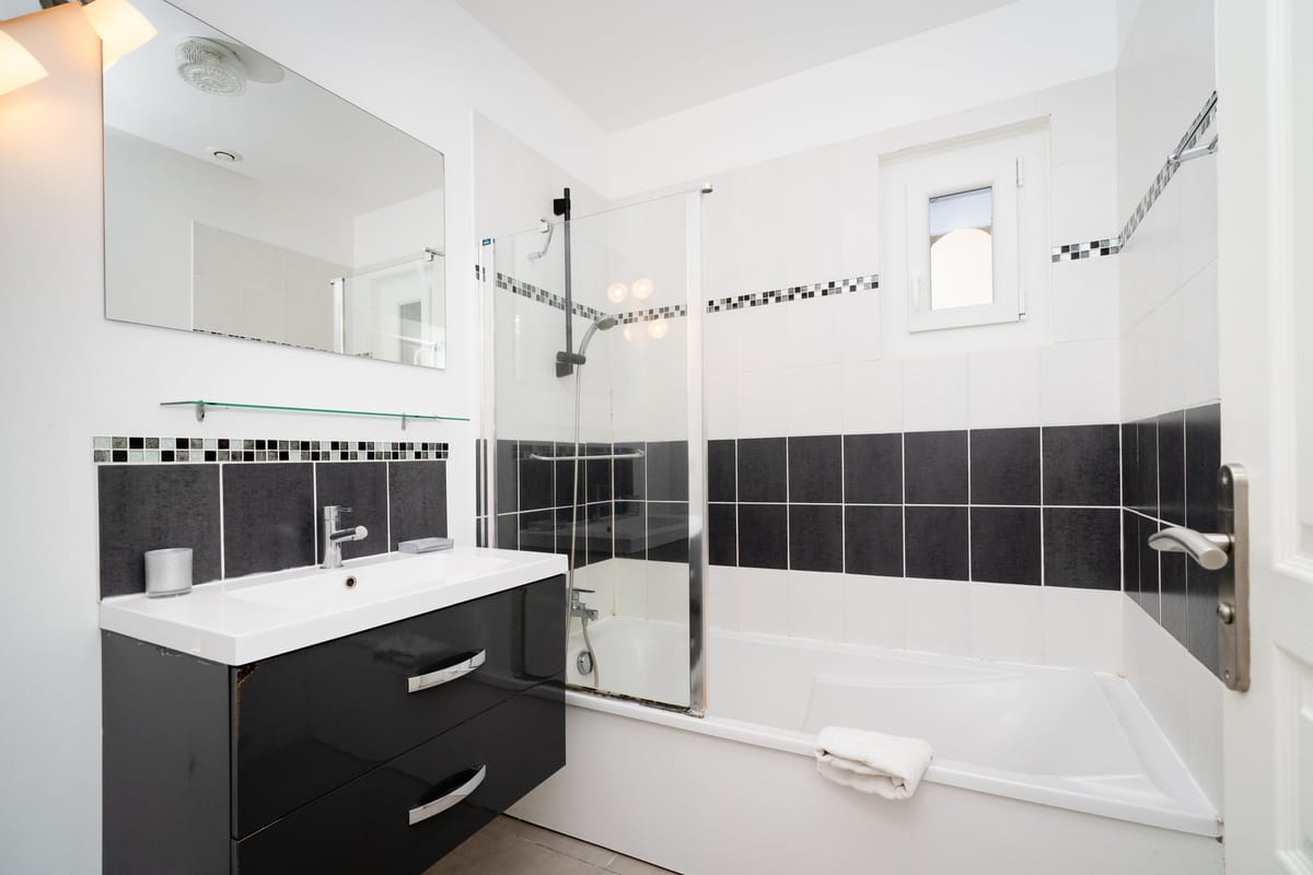 The bathroom of the Les Oliviers gite, with a bath and shower screen, a dark vanity with a basin and a wide mirror.
