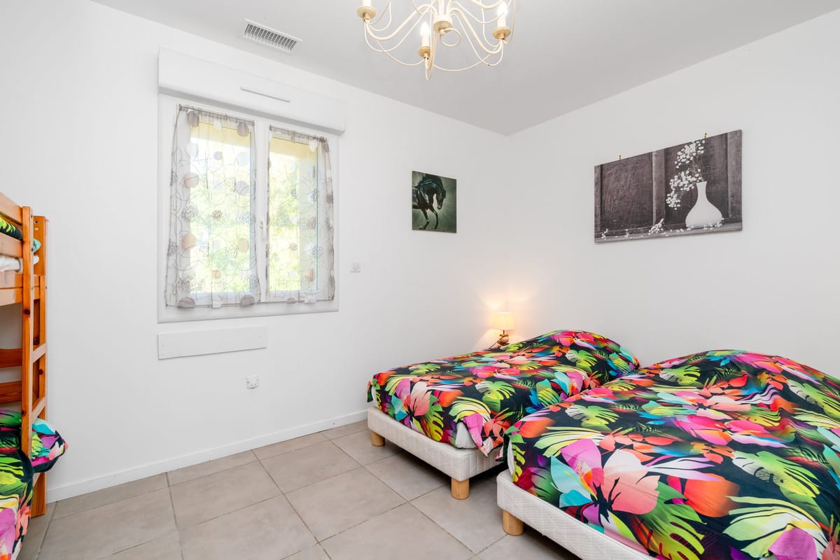 A bedroom in the Les Oliviers gite with two single beds in bright printed covers, a bunk bed and a window onto the trees.