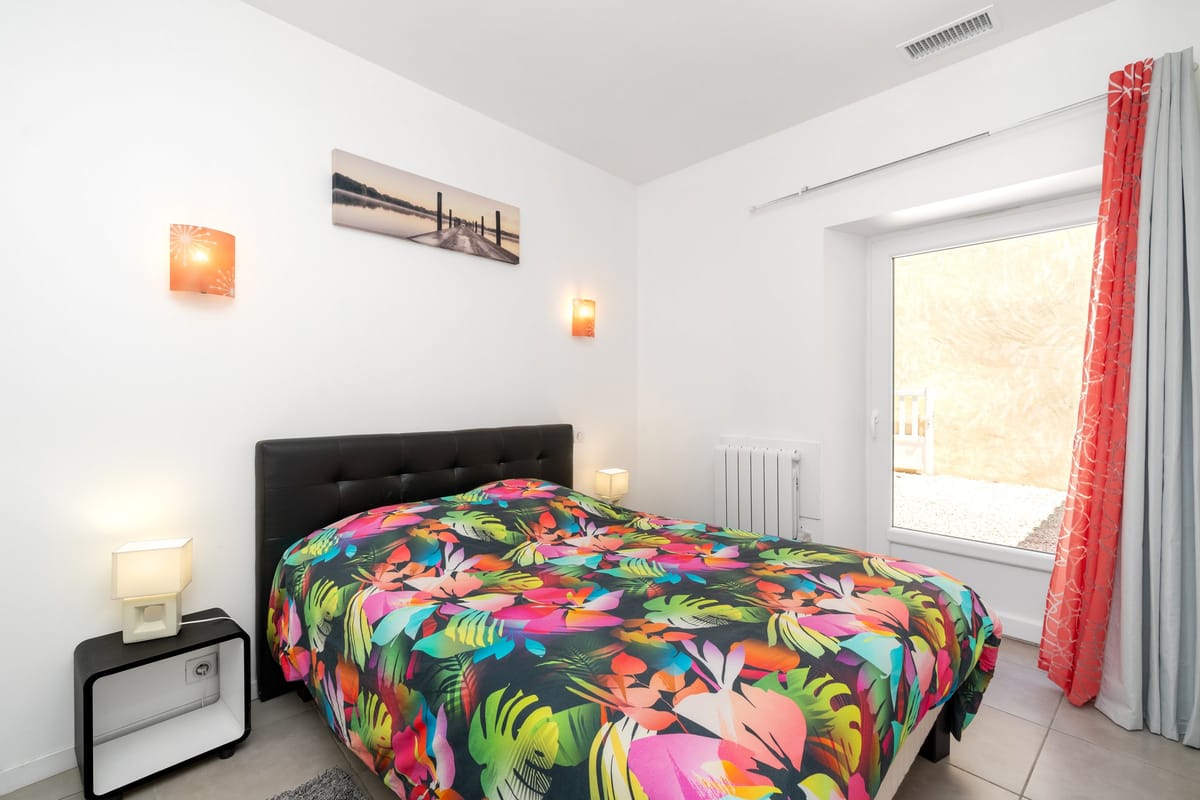 A bedroom in the Les Oliviers gite with a double bed, a black upholstered headboard, wall lights and a window onto the garden.