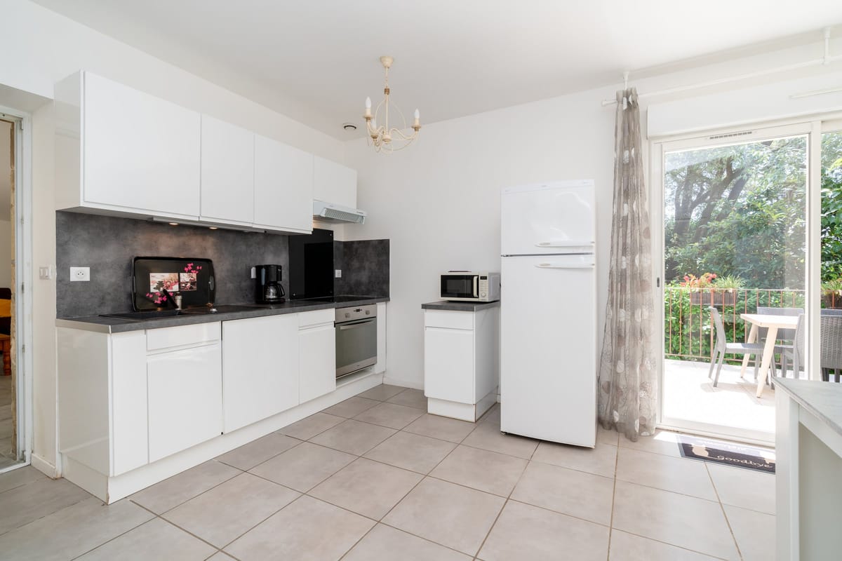 The fitted kitchen of the Les Oliviers gite, with white units, an oven, a fridge and a glazed door onto the terrace.