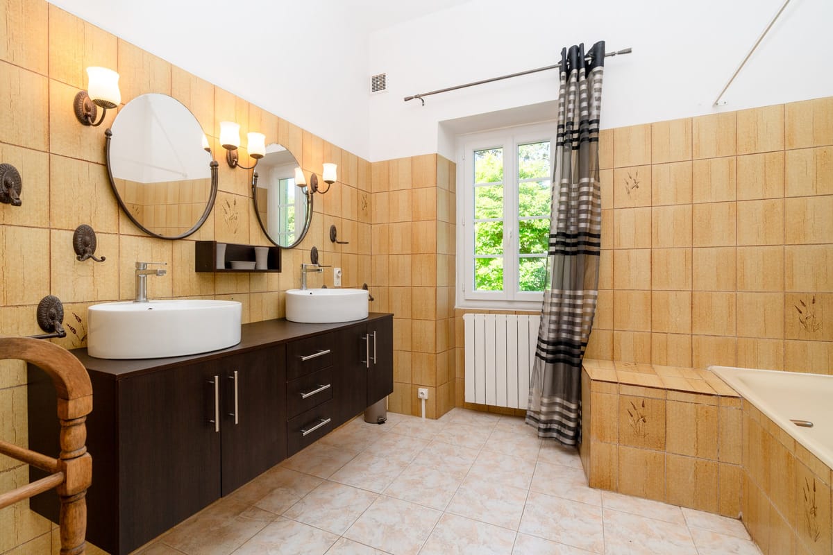 The bathroom of the Les Pins gite, with twin basins on a dark vanity, two round mirrors and ochre tiling.