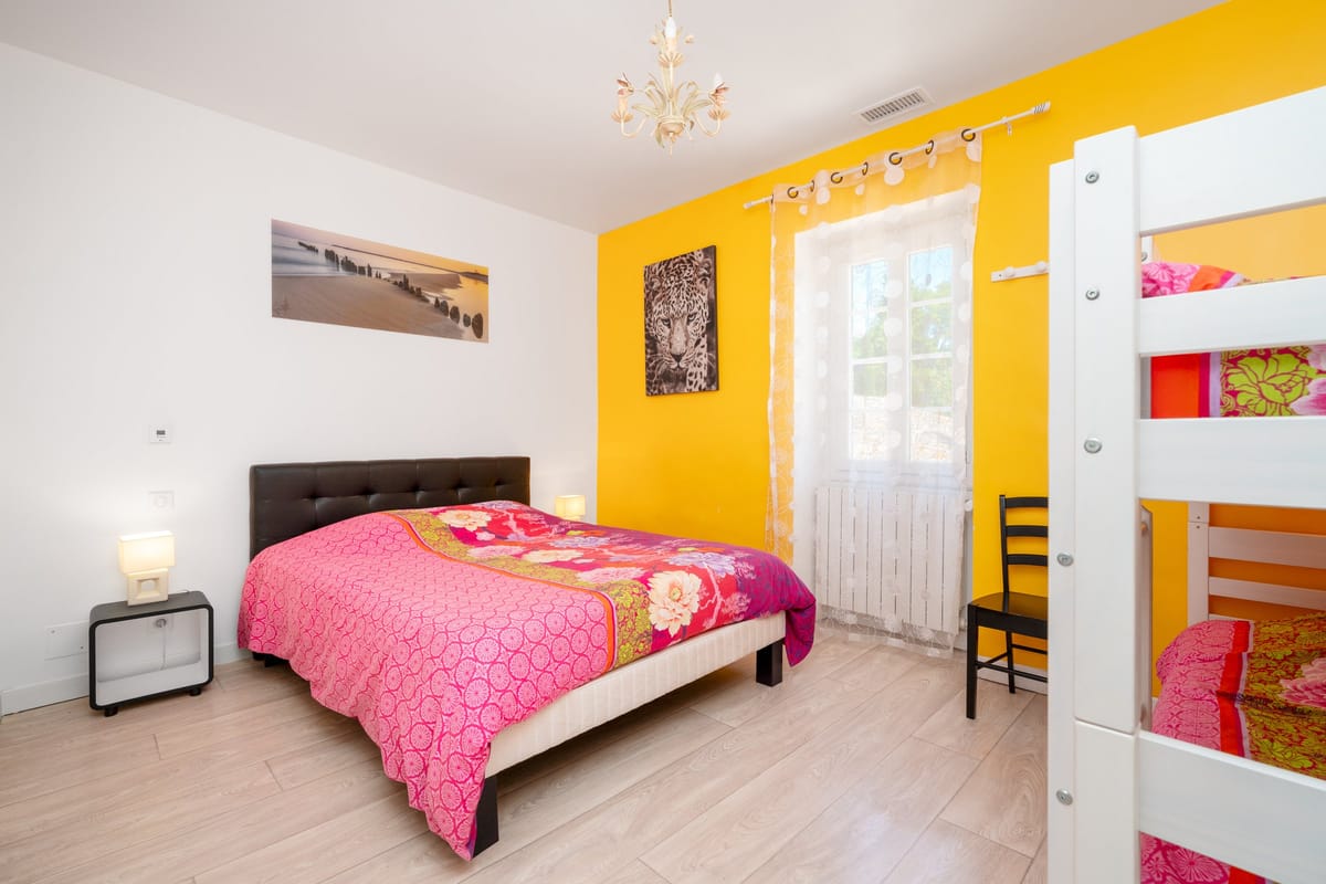 A bedroom in the Les Pins gite with a double bed, a yellow feature wall and a bunk bed alongside.