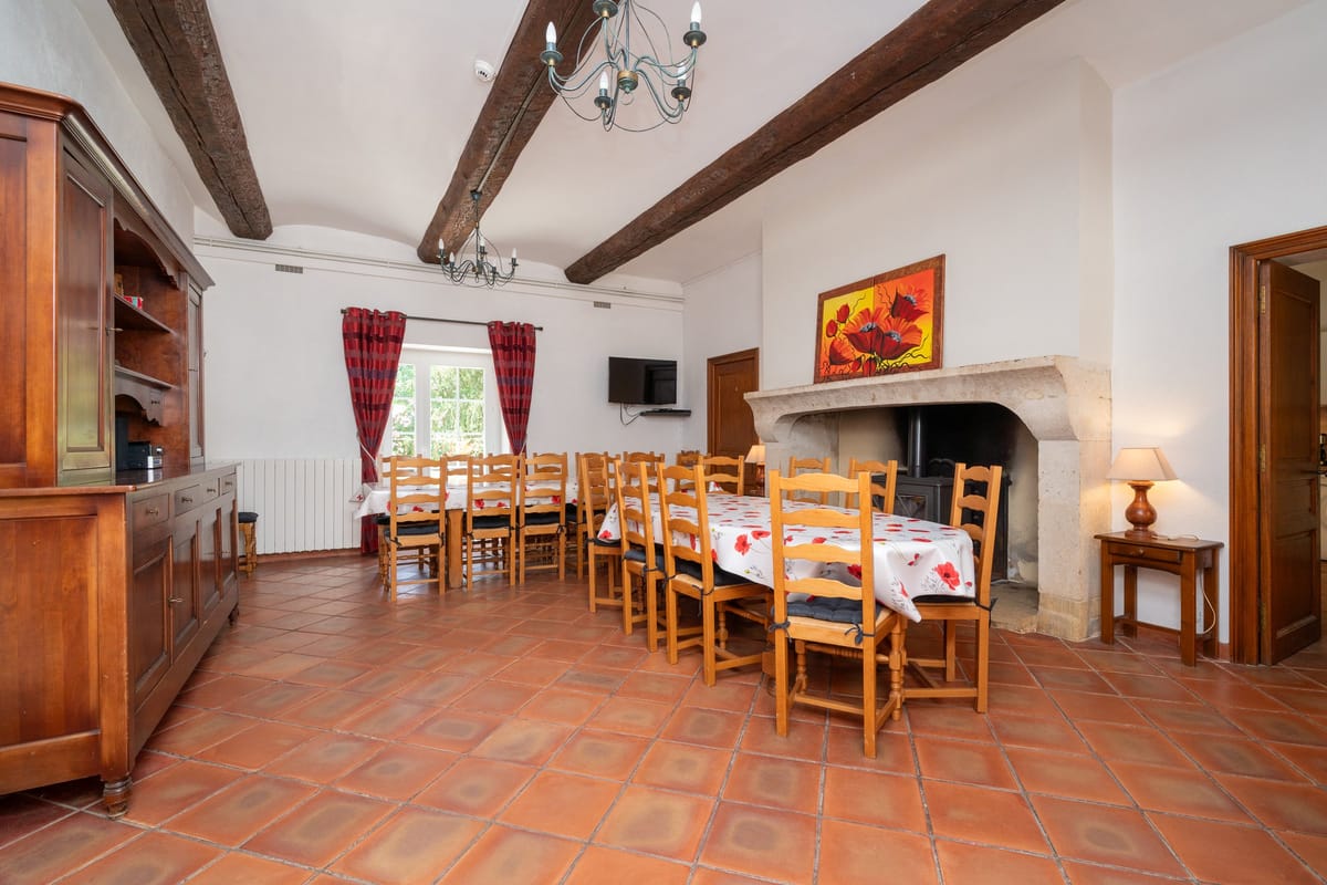 The dining room of the Les Pins gite, with a long table and ladder-back chairs, a stone fireplace, exposed beams and a terracotta tiled floor.