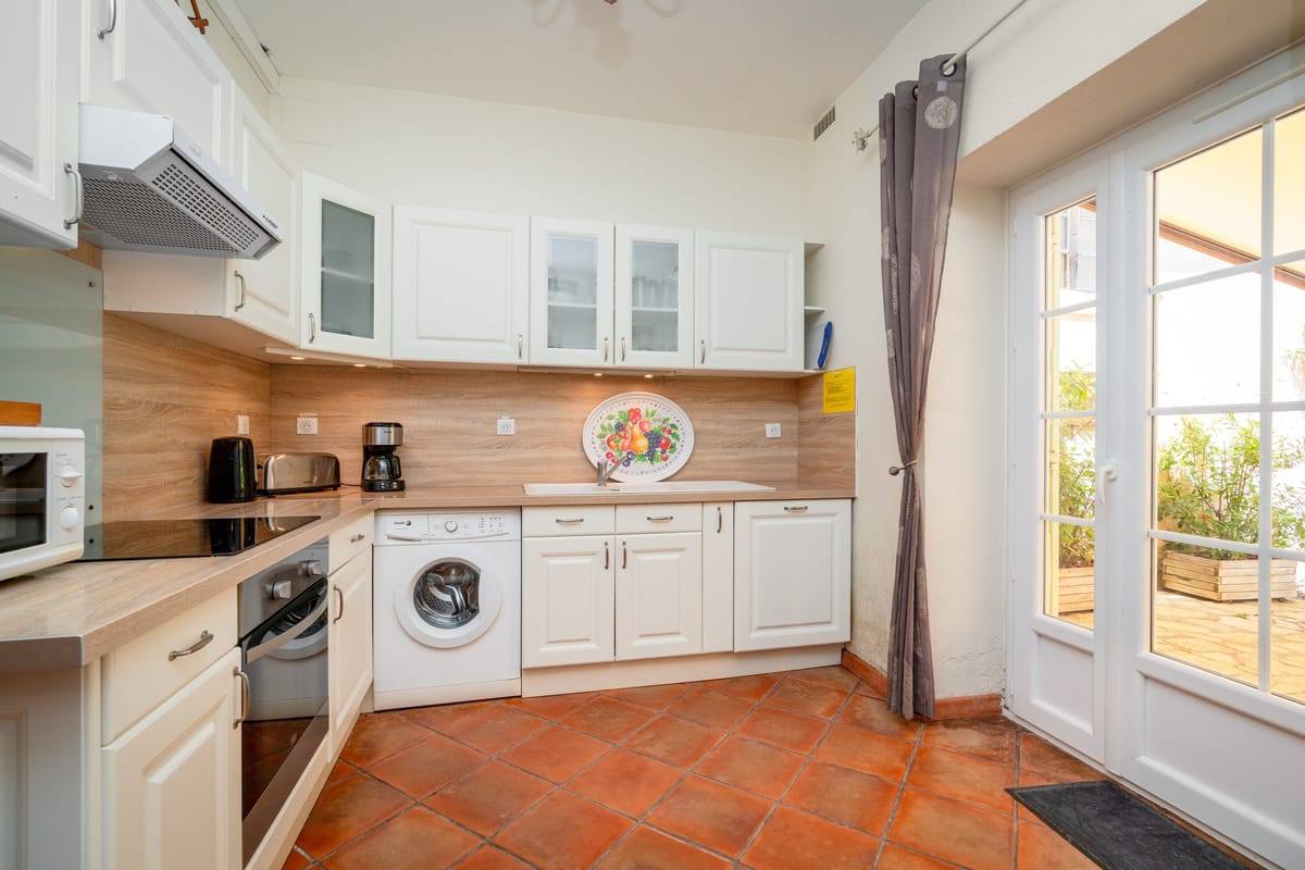 The fitted kitchen of the Les Pins gite, with cream units, a wooden worktop, a washing machine and a glazed door onto the terrace.
