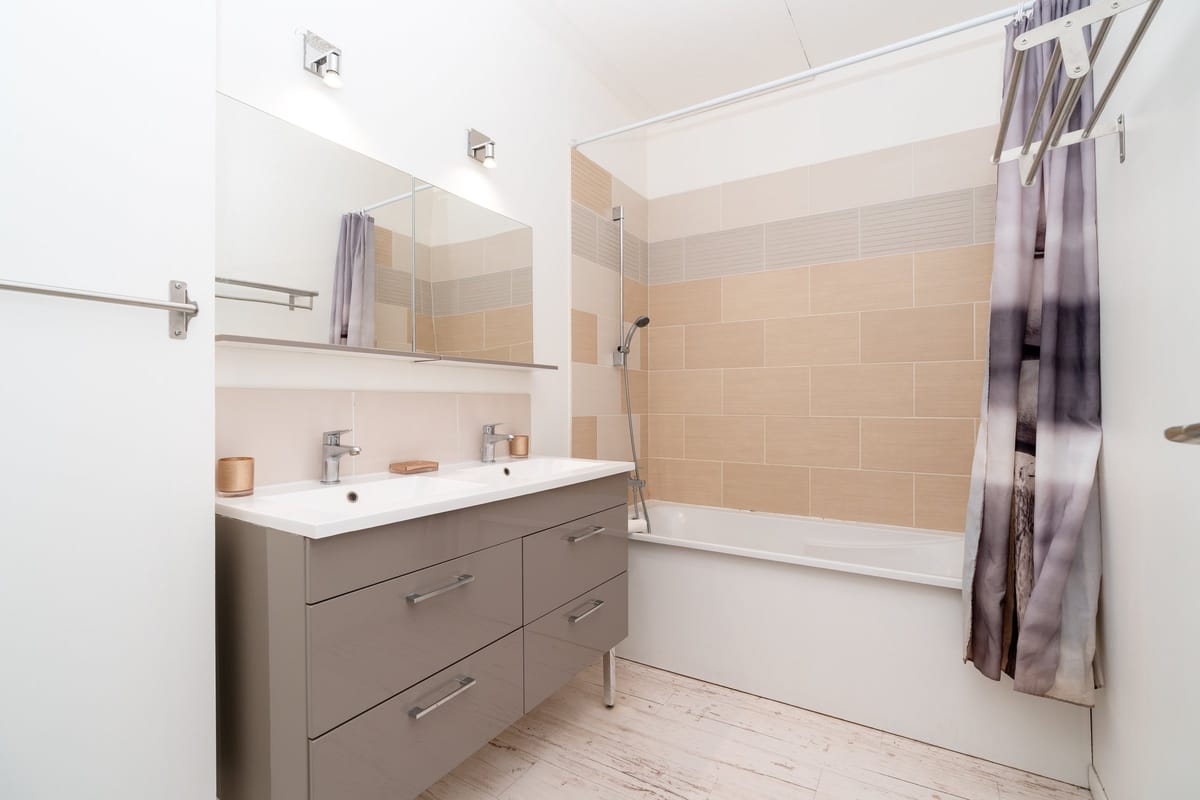 A bathroom in the Les Vignes gite, with twin basins on a taupe vanity and a bath with a shower.