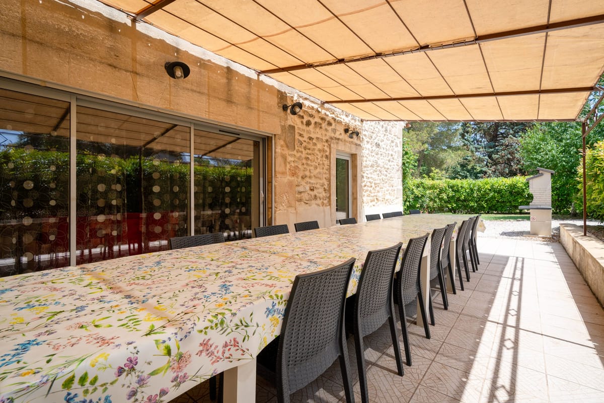 The covered terrace of the Les Vignes gite, laid with a long table and chairs beside the stone building, with the garden and barbecue beyond.