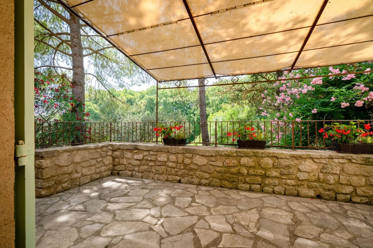 Covered stone terrace with a canvas awning on an iron frame, a stone parapet with potted geraniums and climbing roses, looking out over trees.