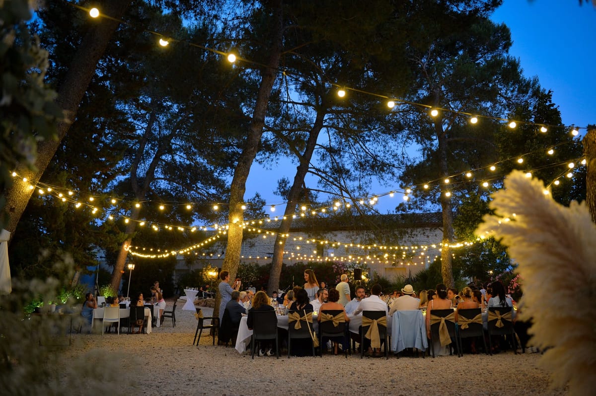 Long tables set on gravel beneath pine trees at dusk, lit by strings of festoon lights, with a tiled building behind.