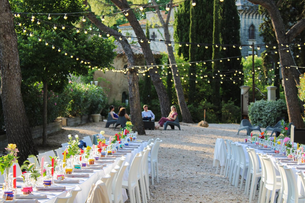 A long banquet table laid on gravel beneath pine trees strung with festoon lights, set with coloured candles and glassware.
