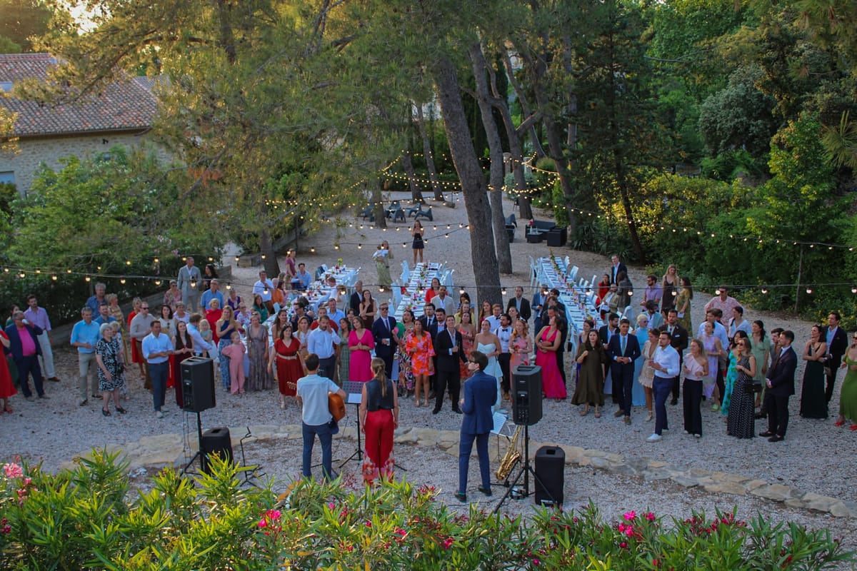 Guests gathered on gravel watching a three-piece band, with long banquet tables and festoon lights among pine trees behind.
