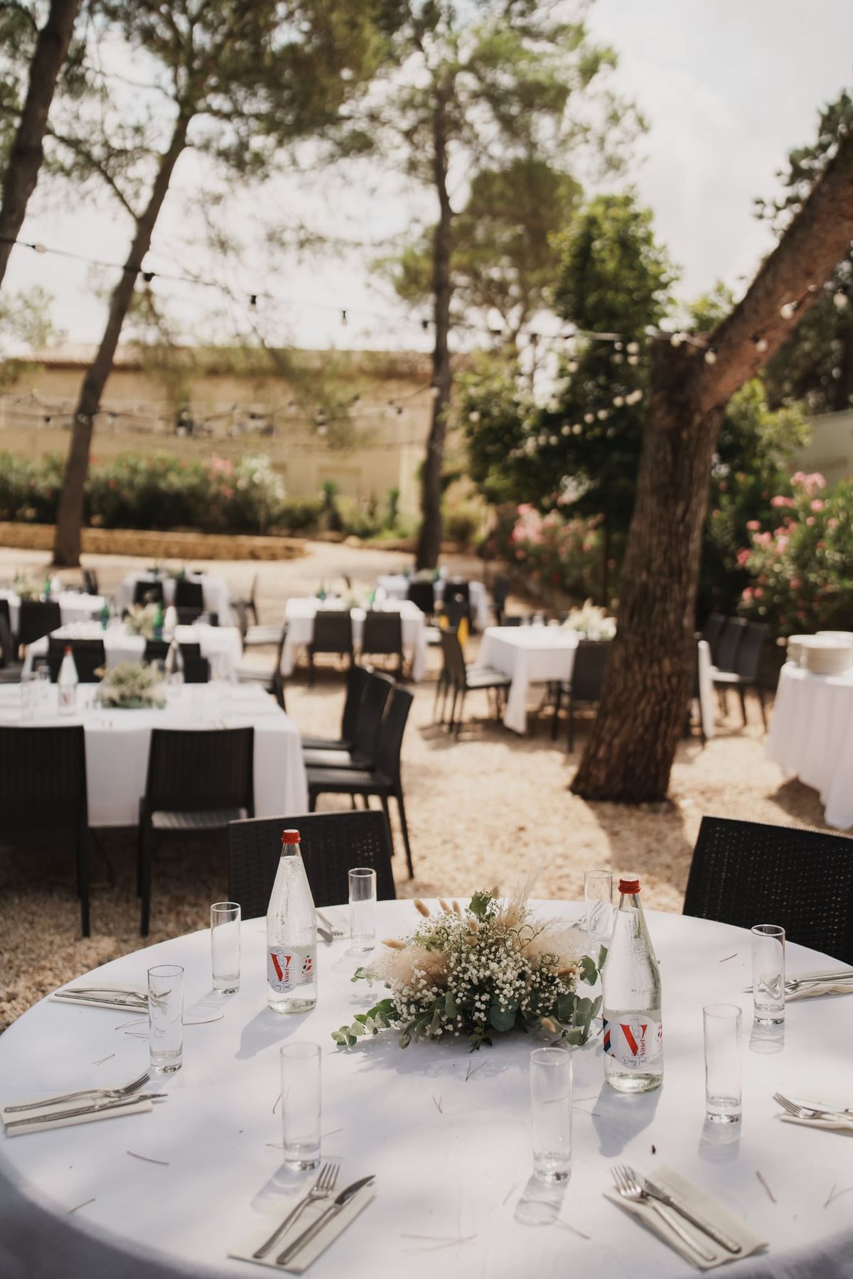 Round table set with white linen, glassware and a posy of gypsophila and pampas, with further set tables on gravel beneath pine trees.