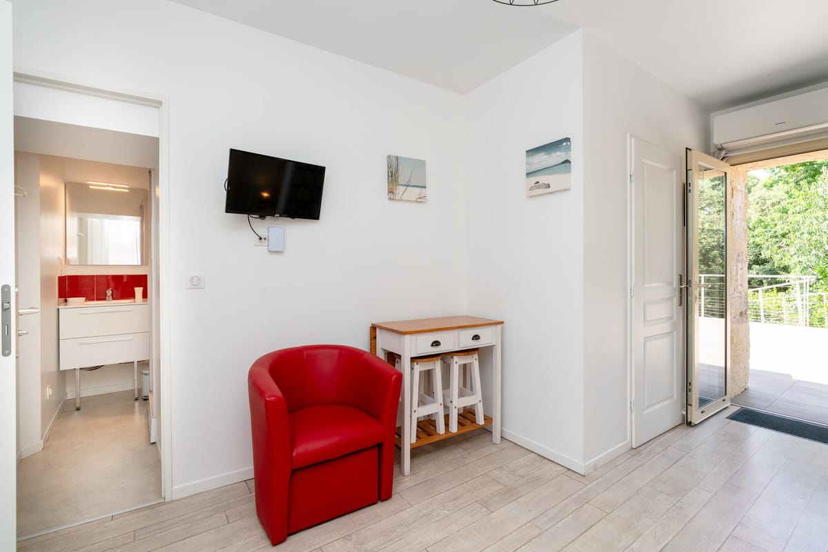 A studio at the estate with a red tub chair, a small table and stools, a wall-mounted television and doors opening onto the terrace.