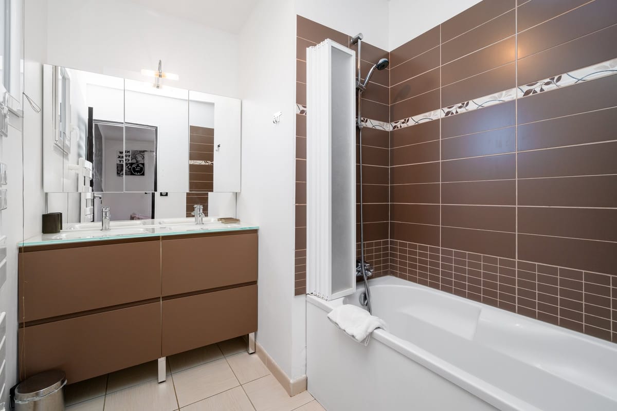 A bathroom in one of the private villas, with twin basins on a brown vanity, a wide mirror and a bath with a shower.