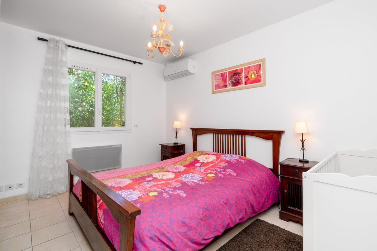 A bedroom in one of the private villas, with a wooden double bed, bedside lamps, a cot and a curtained window.