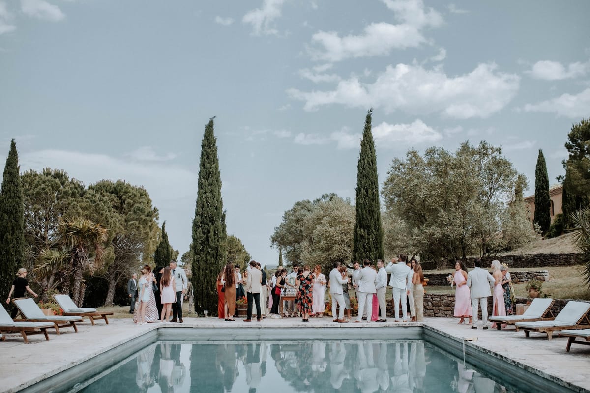 Wedding guests gathered with drinks around the pool, framed by cypress trees and sun loungers