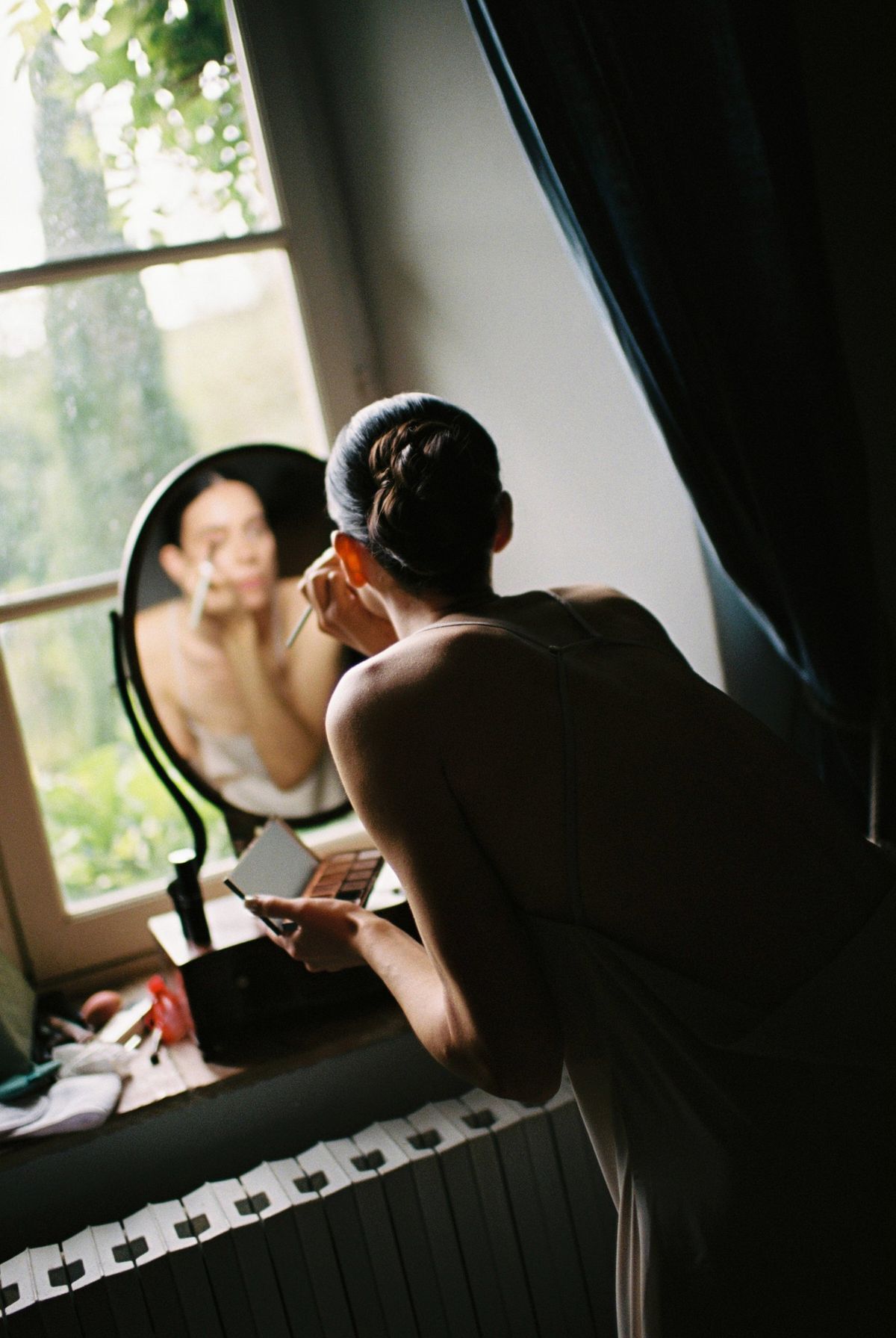 Bride applying make-up at a round mirror on a bedroom windowsill, garden greenery beyond