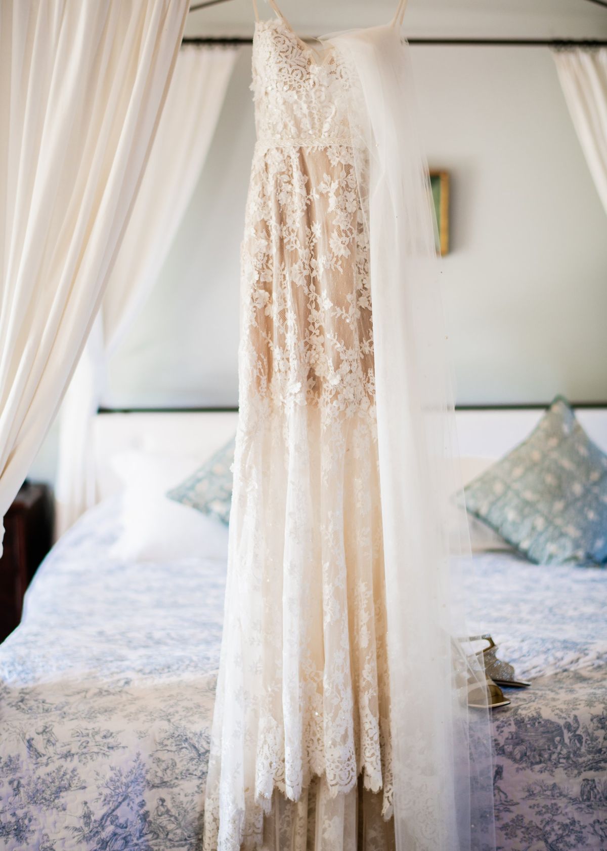 Lace wedding dress and veil hanging from a four-poster bed with toile de Jouy bedding