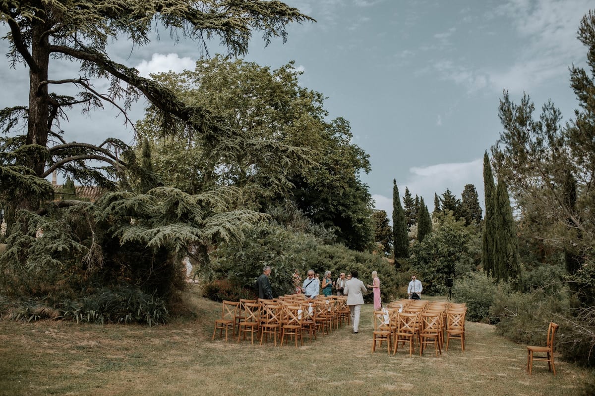 Rows of crossback chairs set for the ceremony on the lawn, guests arriving beneath the trees