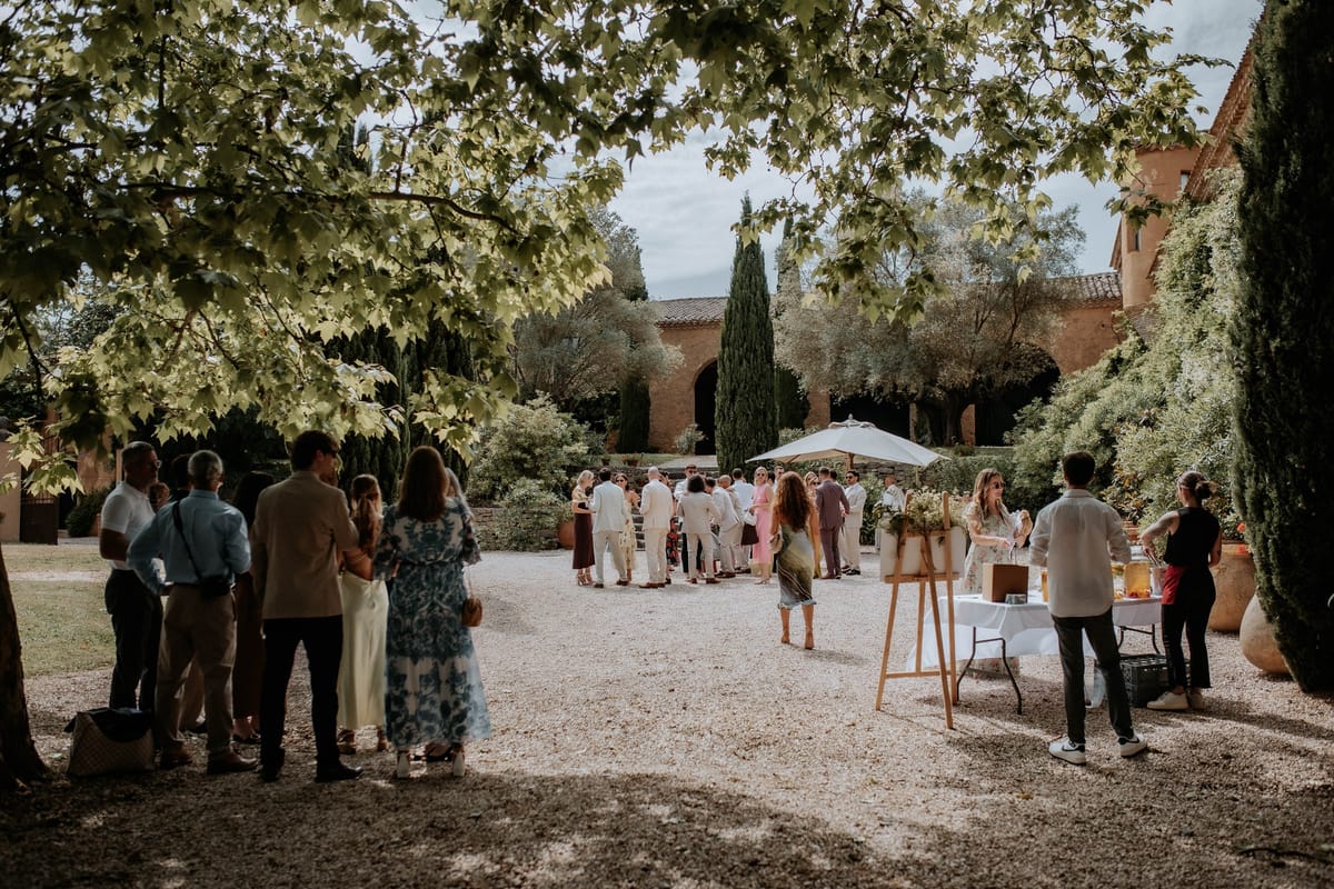 Guests with drinks on the gravel courtyard beneath plane trees, the ochre buildings behind