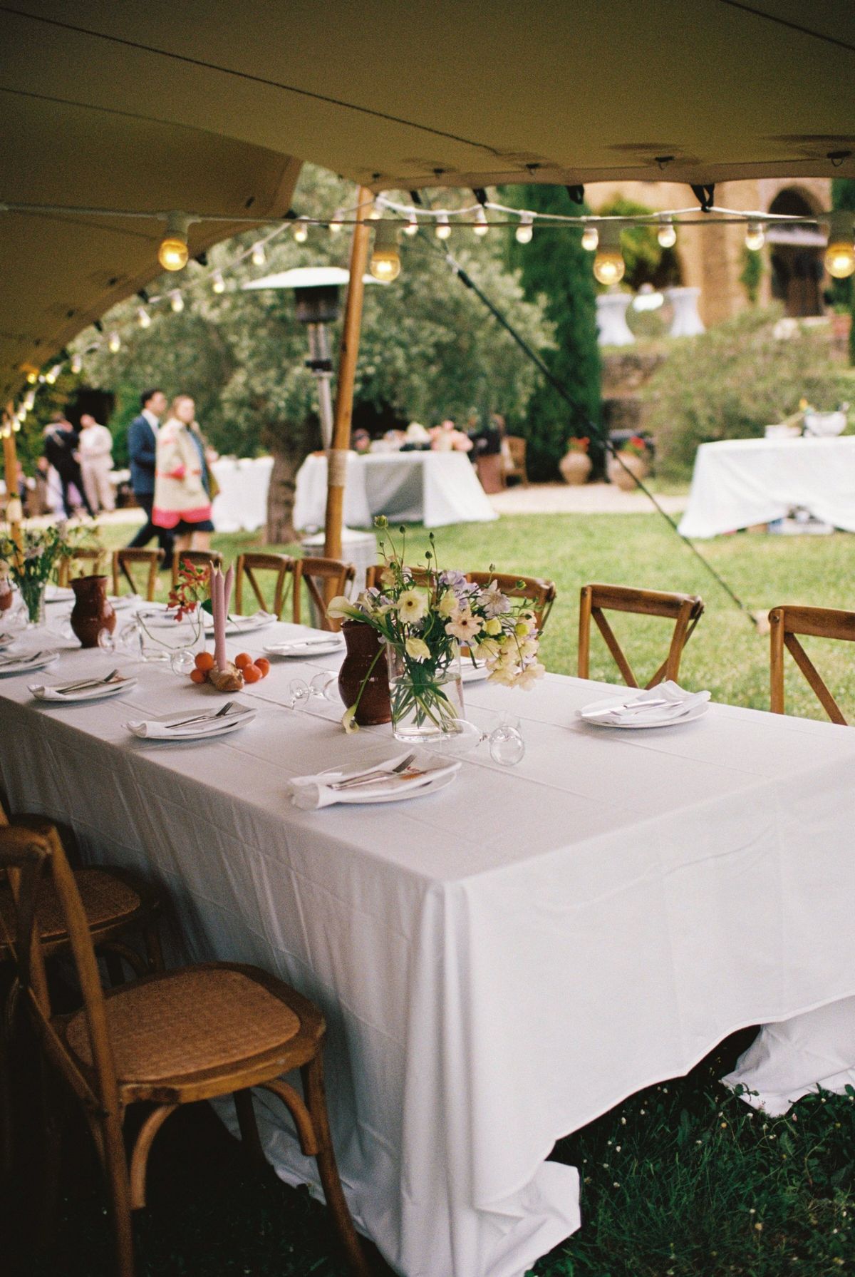Long table under the marquee laid with wildflower posies, festoon lights strung above