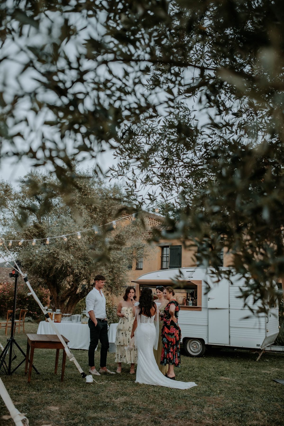 The bride and her guests at the vintage caravan bar at dusk, seen through the olive trees