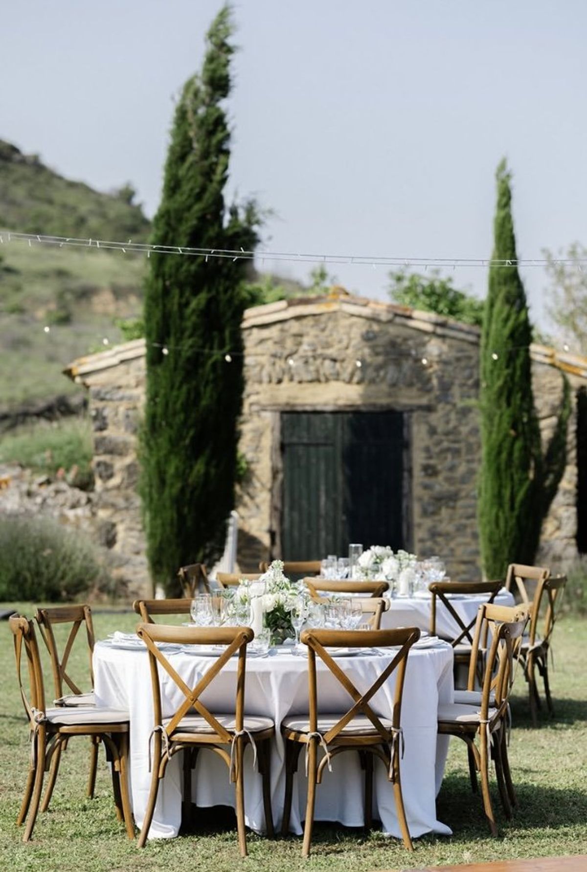 Round table laid with white linen and crossback chairs in front of the stone barn