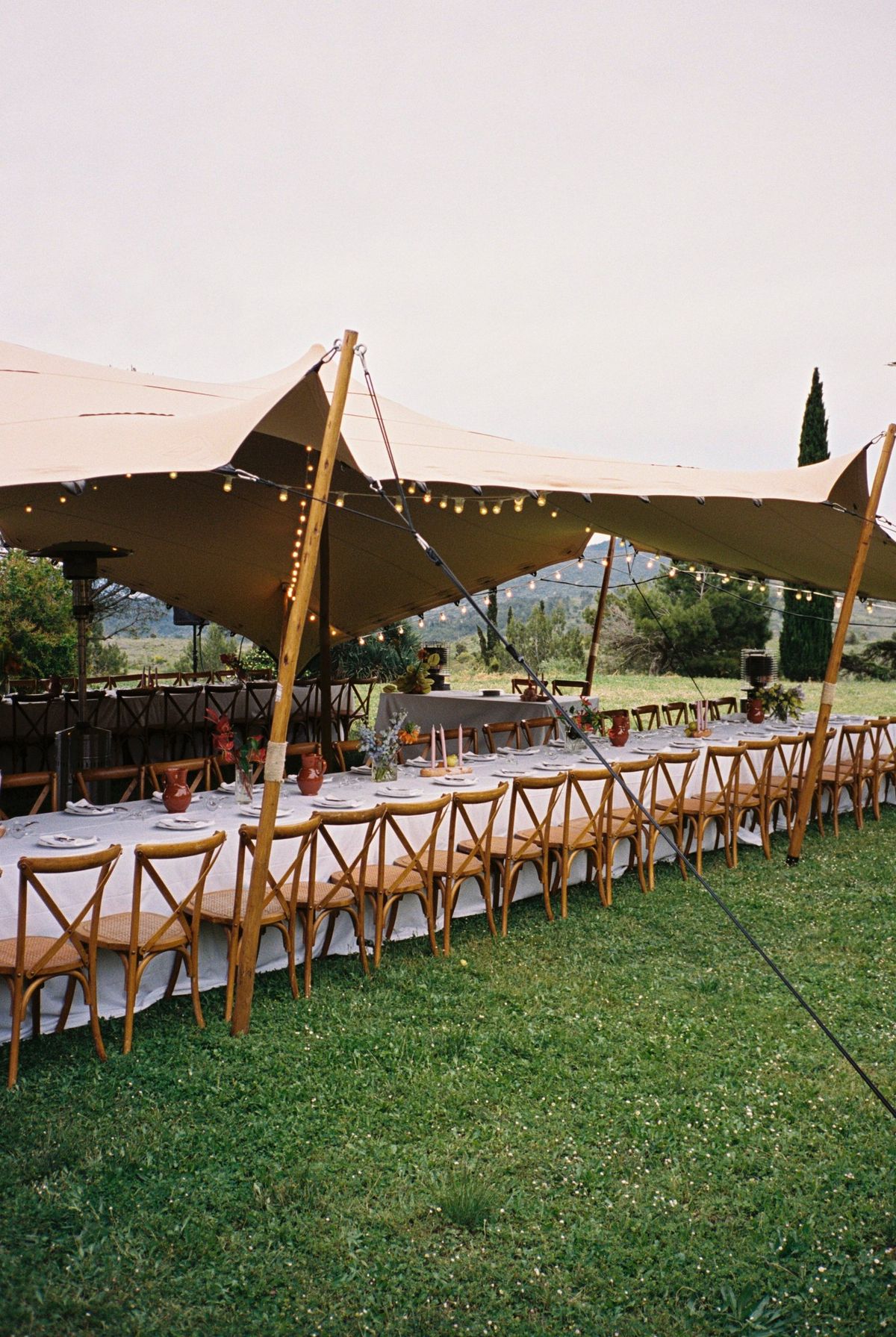 A long banquet table set beneath the stretch tent, festoon lights strung between the poles