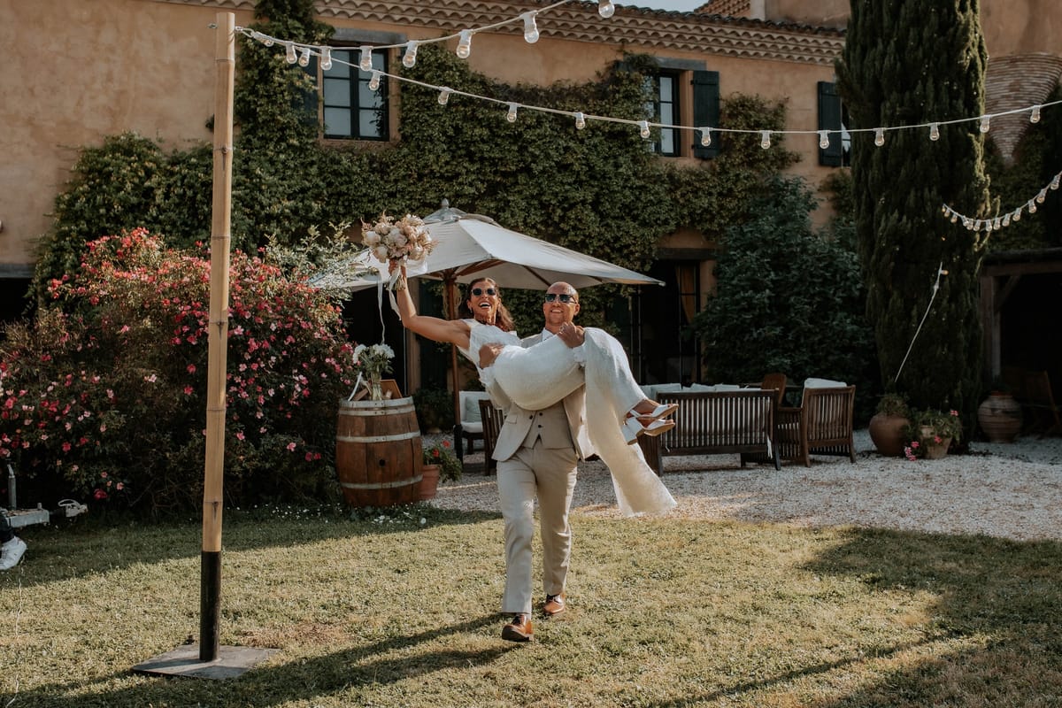 Groom carries the bride across the gravel courtyard beneath strings of festoon lights