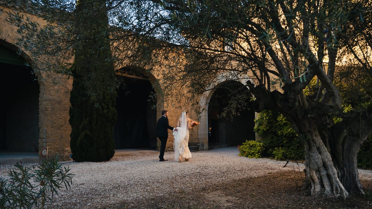 The couple walk together past the stone arches at dusk, beside an old olive tree